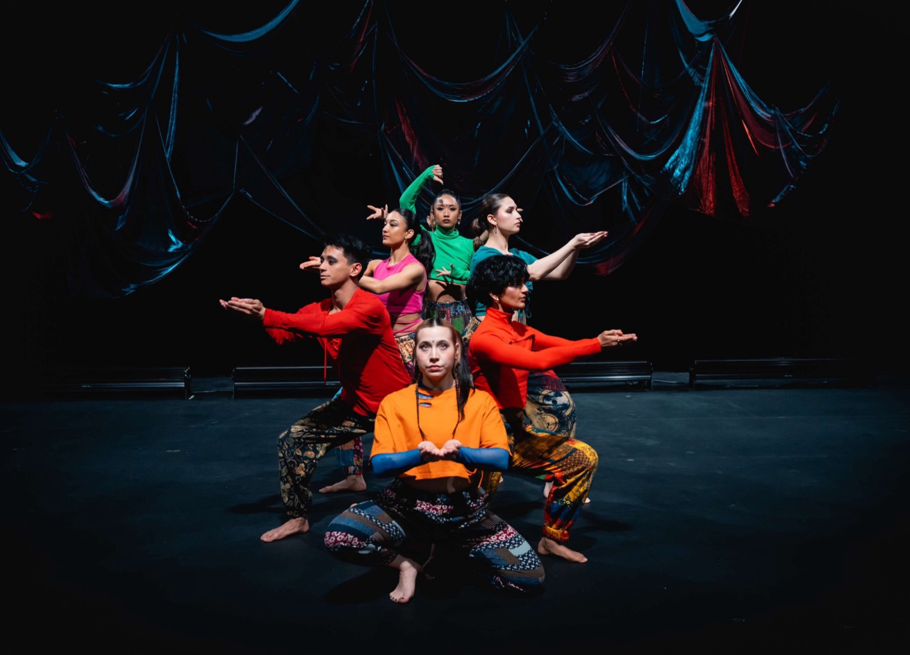 IMGE Dance ensemble in colourful costumes posed on a dark stage